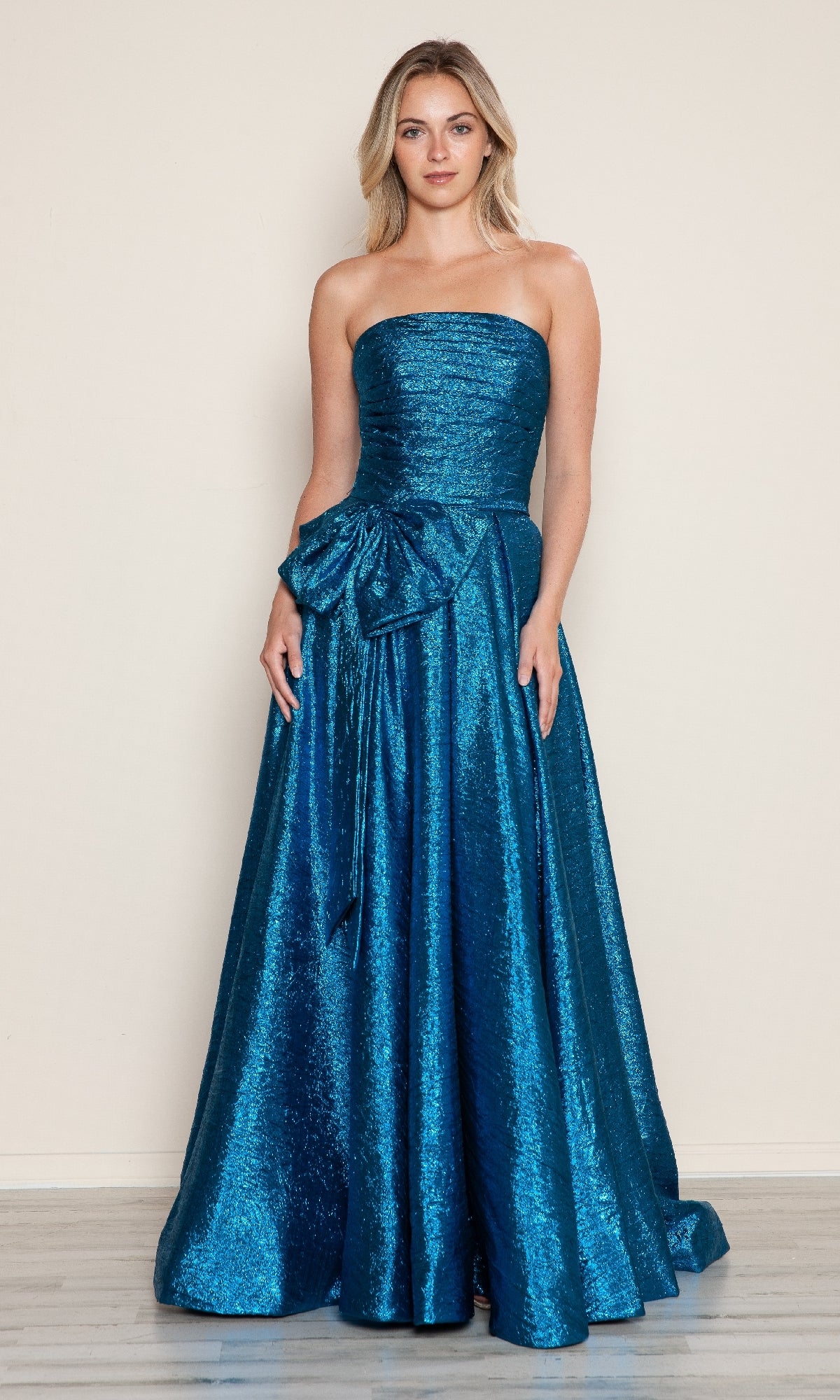 A woman stands against a plain background wearing the Side-Bow Strapless Prom Gown: Poly USA 9720—a floor-length, peacock blue evening dress with a large side bow at the waist.